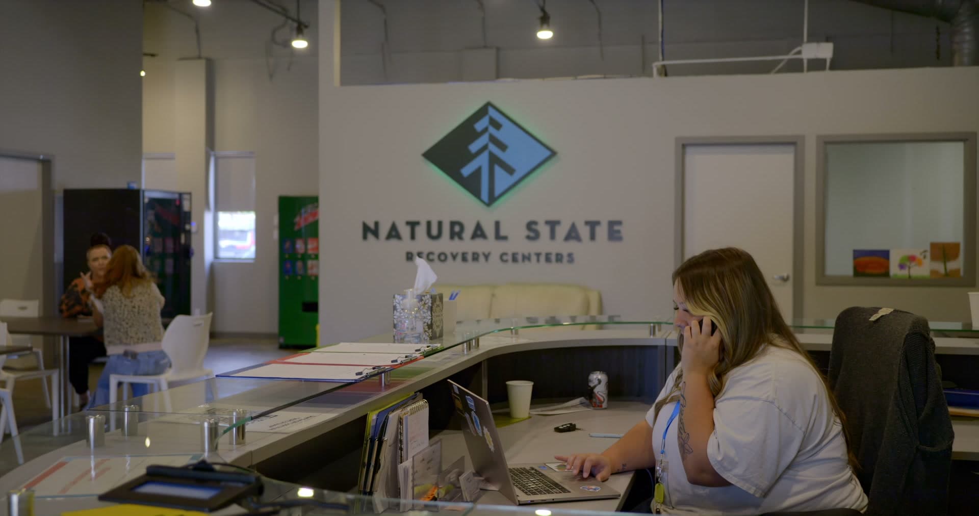 Front desk staff member answering the phone beneath the Natural State Recovery Centers logo