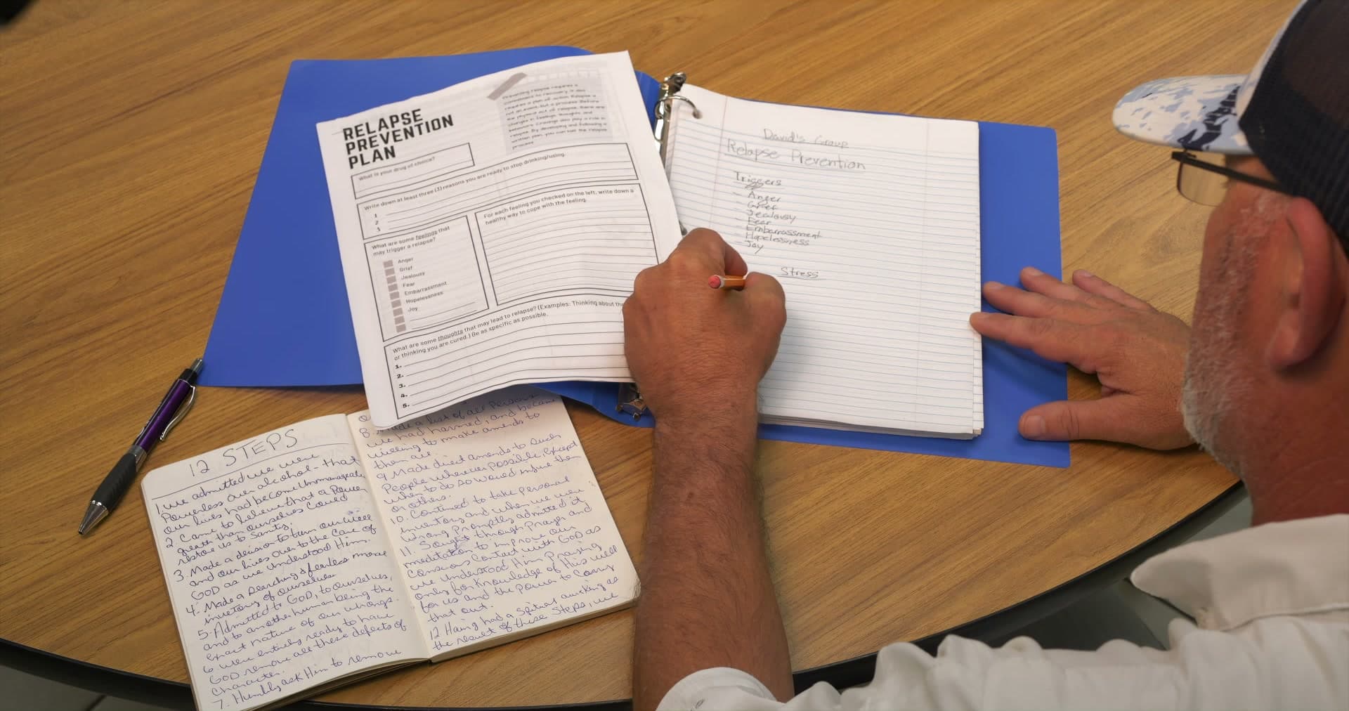 Hand writing a relapse prevention plan beside a notebook with the 12 Steps written out